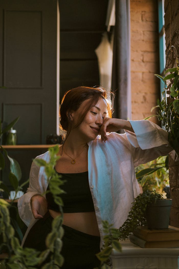A woman in a white shirt enjoys the natural light and indoor plants, exuding a calm and serene mood.