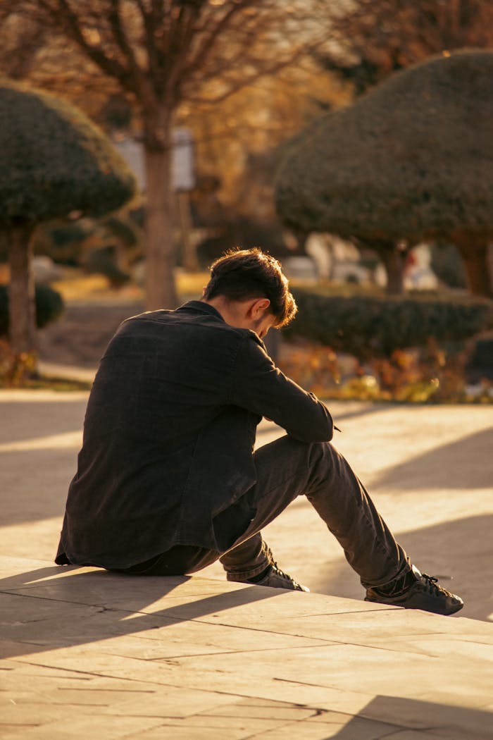 A young man sits alone in a park, deep in thought, during a sunny autumn day.