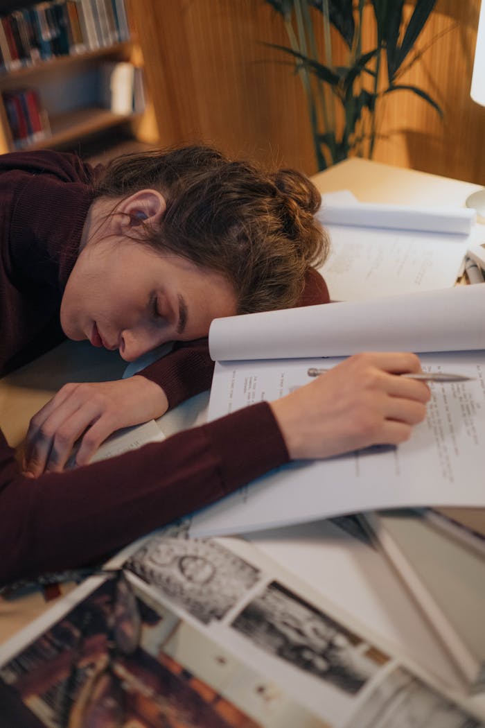 Young woman asleep on a desk surrounded by textbooks and papers, highlighting study exhaustion.